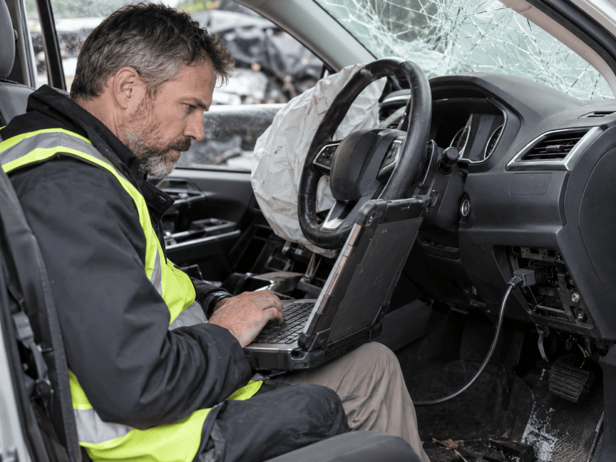 Forensic engineer using a diagnostic laptop connected to a damaged vehicle to retrieve electronic crash data after a road collision.